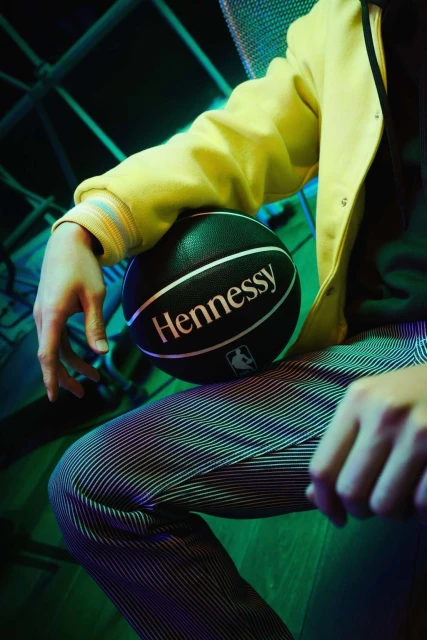 Person sits with a branded Hennessy basketball, wearing a yellow jacket and striped pants, under cool green and blue lighting.