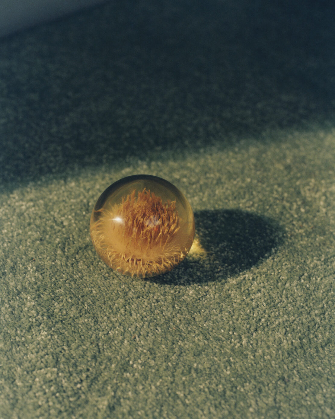 An amber-colored glass ball with organic filament-like inclusions sits on a soft, green carpet in soft, natural lighting. 