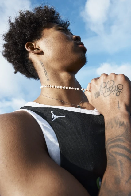 Young adult male in black athletic top glances upward in a medium shot, with a pearl necklace, tattoos, and a cloudy blue sky.