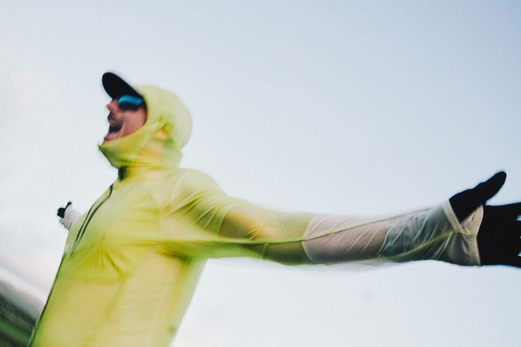 A man in a translucent yellow waterproof hooded jacket cheering with arms outstretched against an open sky.