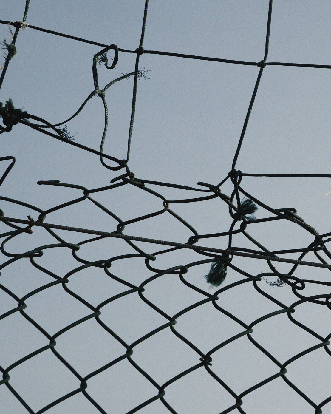 Close-up of a damaged chain-link fence against a pale blue sky, showing signs of wear and tear and a hole in the netting.