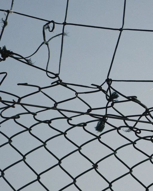 Close-up of a damaged chain-link fence against a pale blue sky, showing signs of wear and tear and a hole in the netting.
