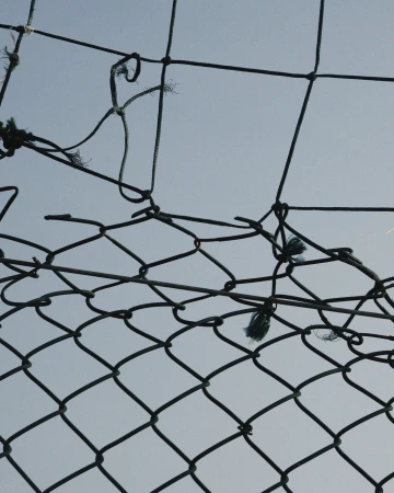 Close-up of a damaged chain-link fence against a pale blue sky, showing signs of wear and tear and a hole in the netting.