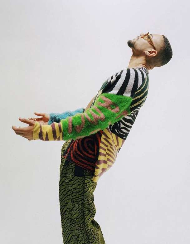 A man with tan skin, wearing a brightly patterned sweater and glasses, stands in front of a solid white backdrop in a studio setting.