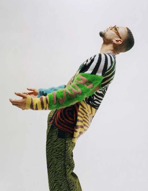 A man with tan skin, wearing a brightly patterned sweater and glasses, stands in front of a solid white backdrop in a studio setting.
