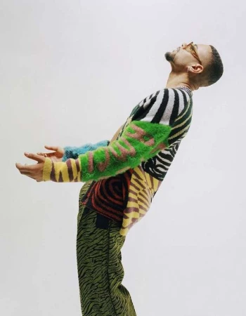 A man with tan skin, wearing a brightly patterned sweater and glasses, stands in front of a solid white backdrop in a studio setting.