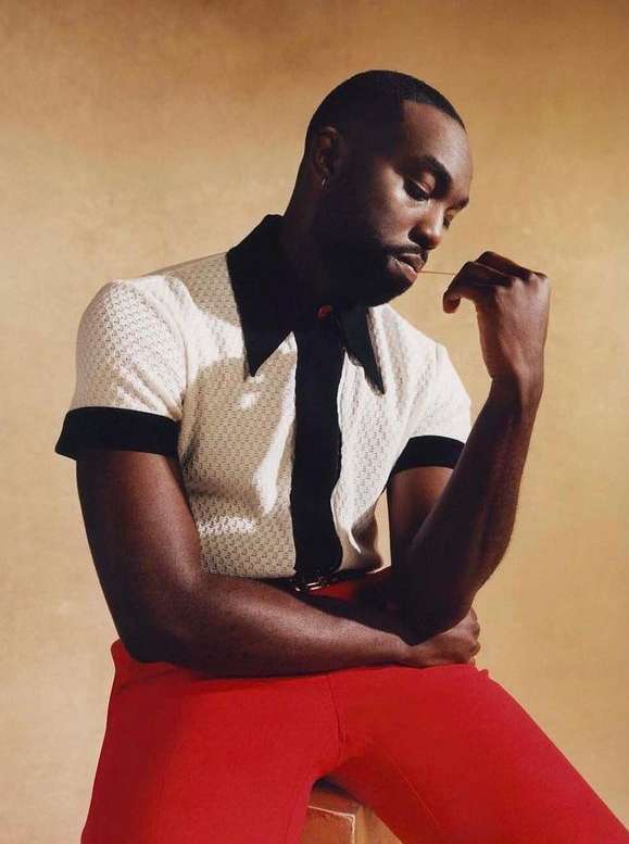 Portrait of a dark-skinned adult male wearing a white and black polo shirt with red pants, sitting against a tan backdrop.