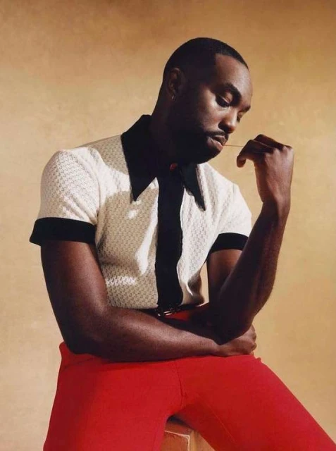 Portrait of a dark-skinned adult male wearing a white and black polo shirt with red pants, sitting against a tan backdrop.