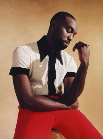 Portrait of a dark-skinned adult male wearing a white and black polo shirt with red pants, sitting against a tan backdrop.