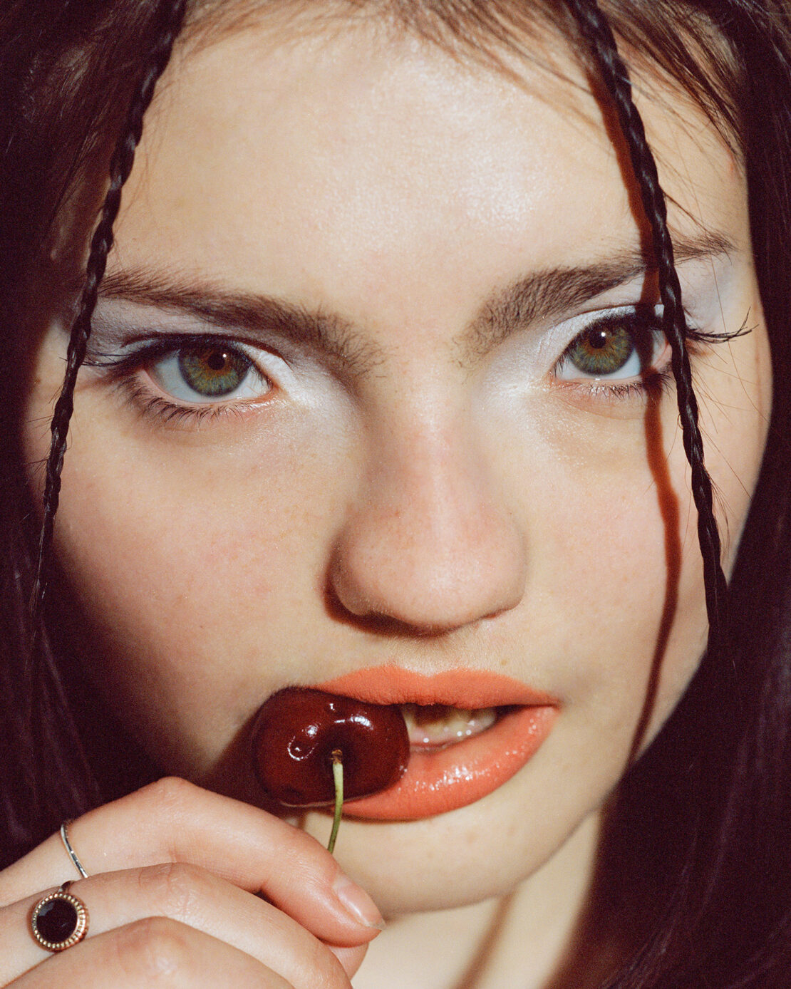 Close-up portrait of a woman biting a cherry, wearing bright coral lipstick and white eye shadow, photographed by Elliott Wilcox.