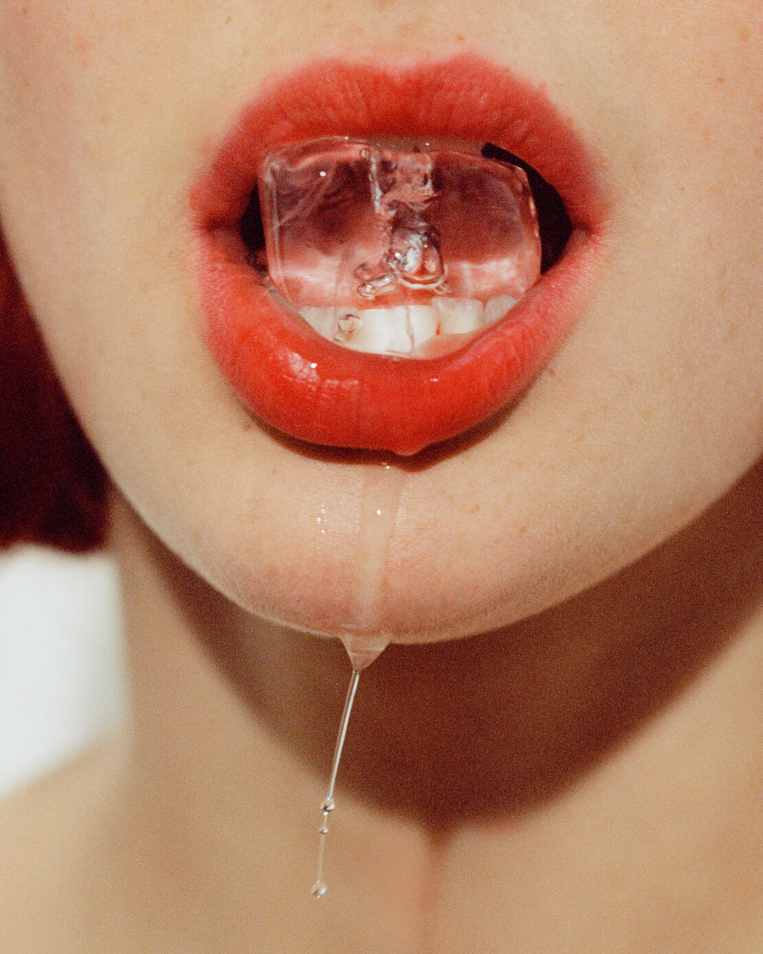 Close-up of a person's mouth with bright red lipstick holding a clear ice cube between their teeth, some melting water droplets.