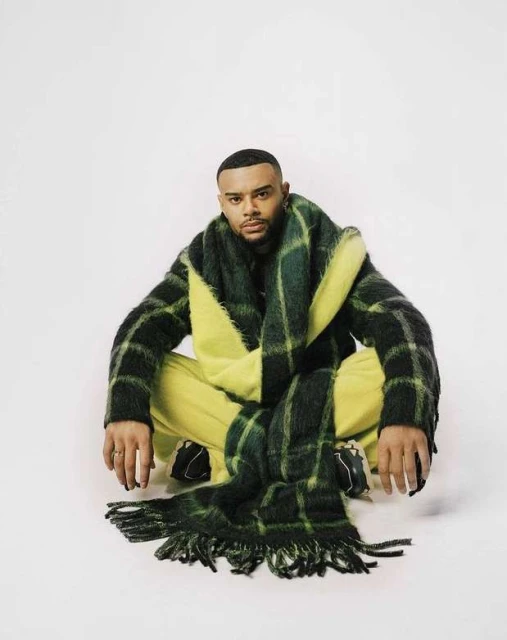 Portrait of a young adult male crouching, wearing yellow pants and a green plaid jacket with a matching fringed scarf on a white backdrop.