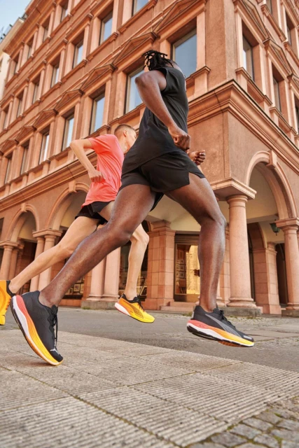 Two athletes run on a city street near a building; one is a Black adult male in black athletic wear, the other is a light-skinned male.