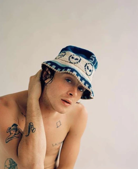 Portrait of a young adult male with a crochet bucket hat, looking at the camera with tattoos, against a neutral studio background.