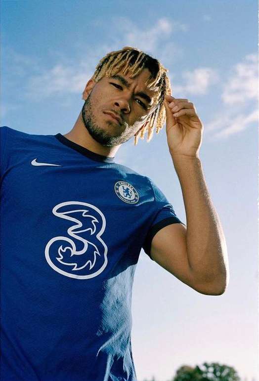 Male soccer player in blue Chelsea jersey with Three logo poses outdoors against blue sky with clouds, shot from a low angle.