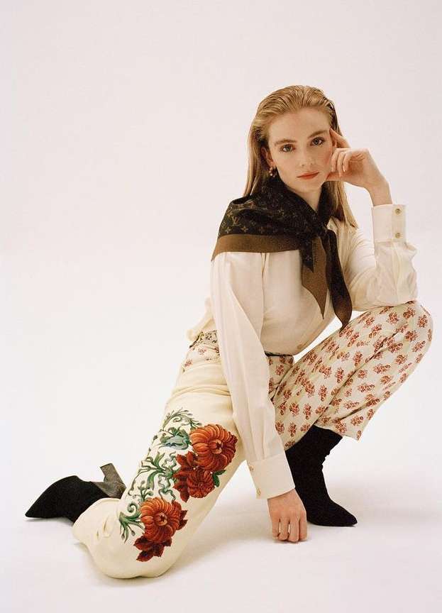 Fashion photo of a woman crouching in a studio wearing a white blouse, floral pants, and boots with a patterned scarf.