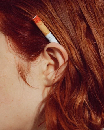 Close-up of a person with vibrant red hair tucking a cigarette behind their ear, photographed by Elliott Wilcox.