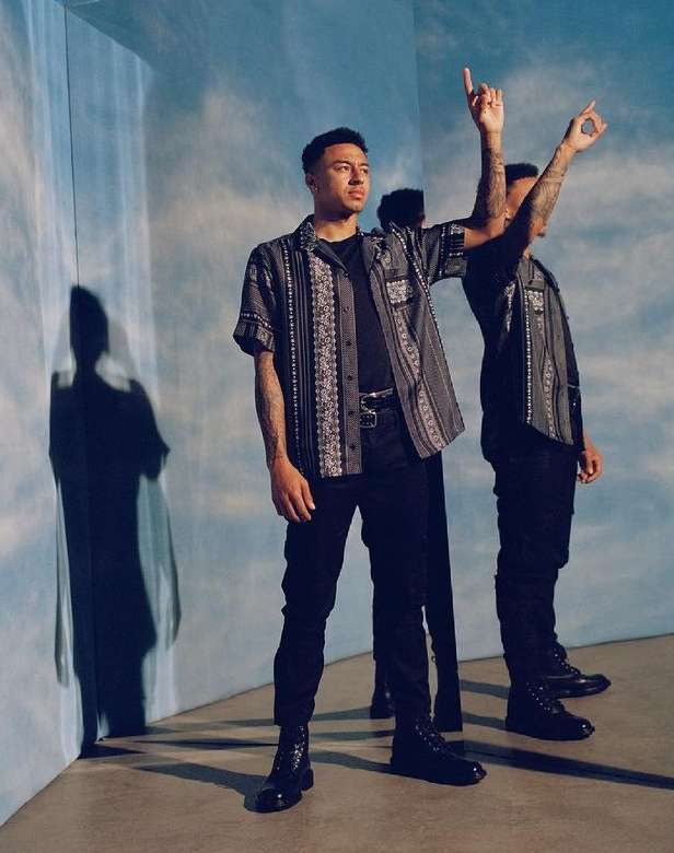 An adult man poses in a patterned shirt and black pants in a studio with a shadow and blurred mirror reflection.
