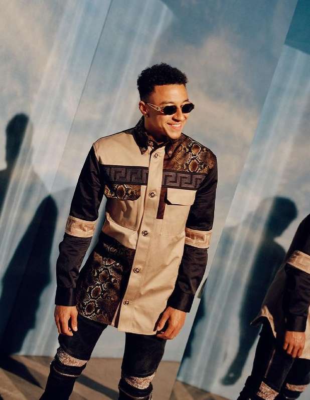 A stylish young man in a designer jacket and sunglasses smiles in a studio setting with a light blue background and mirror reflections.