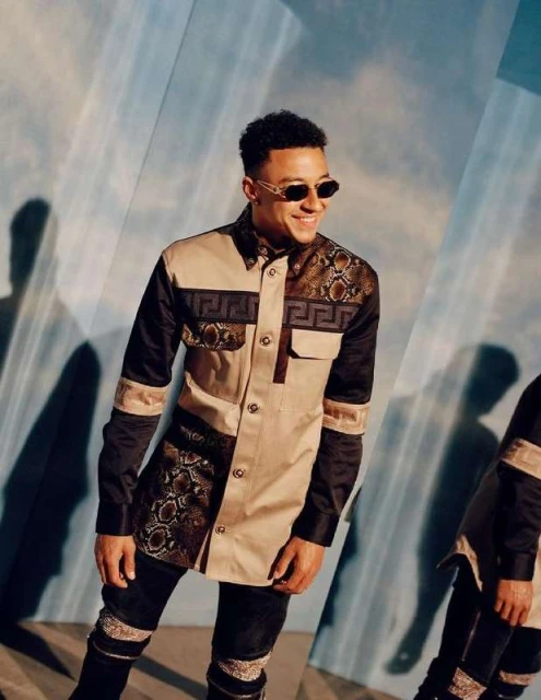 A stylish young man in a designer jacket and sunglasses smiles in a studio setting with a light blue background and mirror reflections.