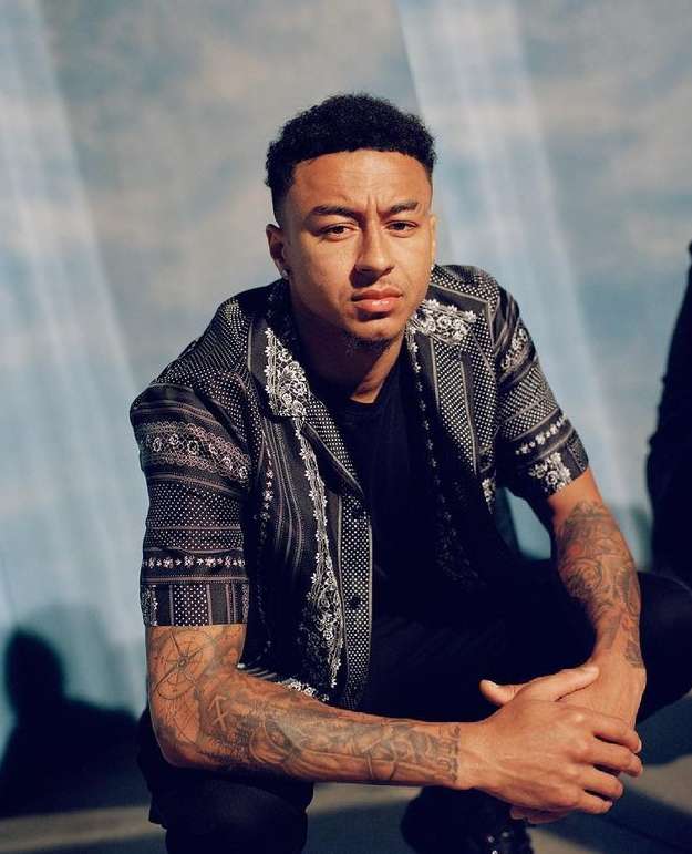Portrait of a man with tattoos on his arm wearing a patterned shirt, close-up shot in front of a blue and white backdrop, eye-level angle.
