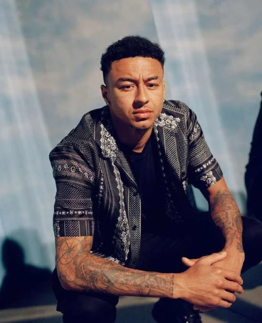 Portrait of a man with tattoos on his arm wearing a patterned shirt, close-up shot in front of a blue and white backdrop, eye-level angle.
