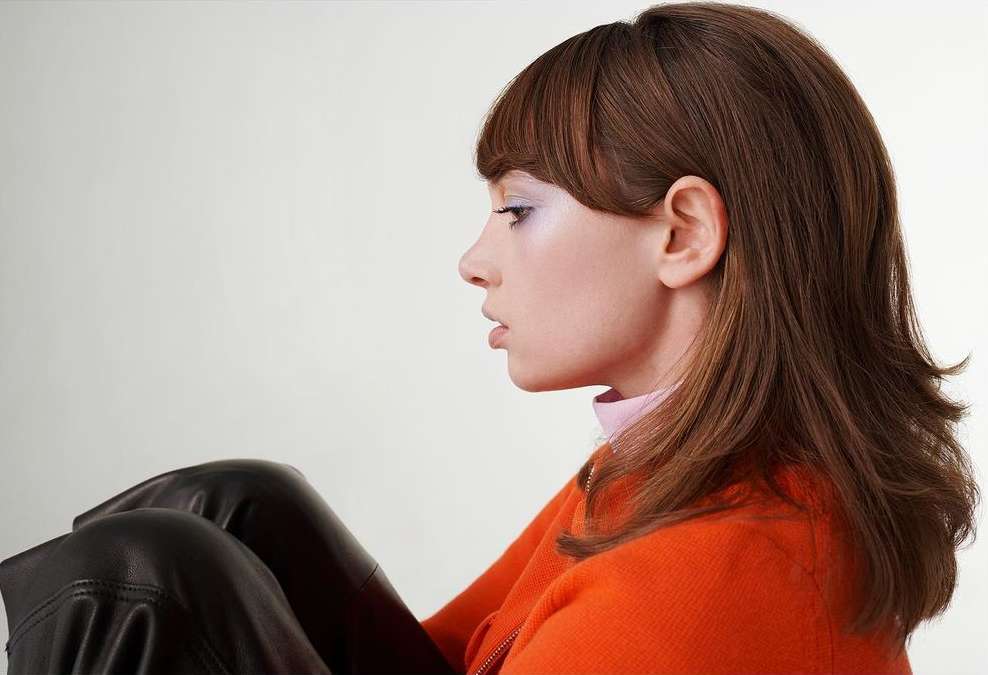 Profile shot of a young woman with brown hair wearing an orange sweater and black leather pants, set against a clean white background.