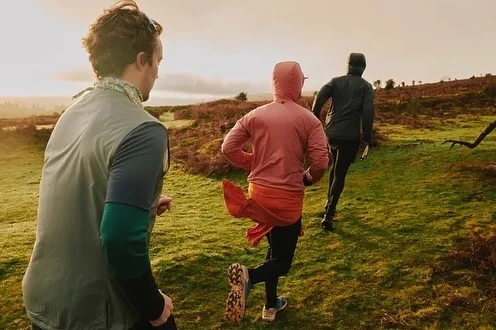 Three people trail running through a grassy field under an overcast sky, wearing athletic clothing on a sunny day.