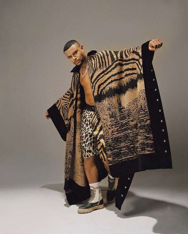 Man in animal print cape poses in photo studio wearing leopard shorts and combat boots against neutral grey backdrop.