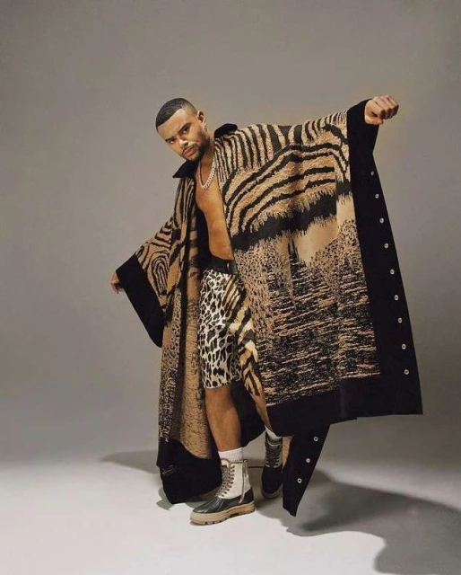 Man in animal print cape poses in photo studio wearing leopard shorts and combat boots against neutral grey backdrop.
