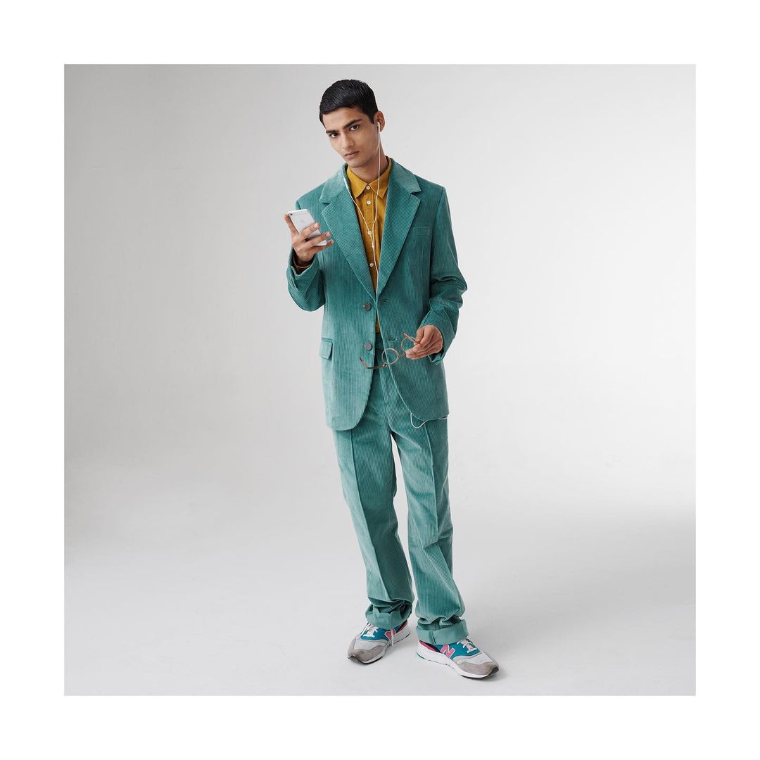 A young man in a green corduroy suit stands in a photo studio looking at a cell phone, with glasses hanging from his shirt.
