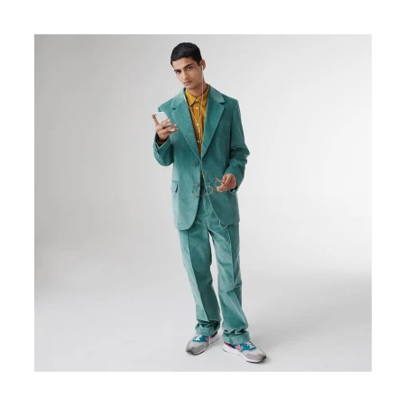 A young man in a green corduroy suit stands in a photo studio looking at a cell phone, with glasses hanging from his shirt.