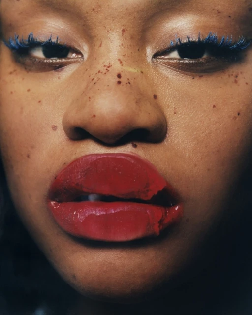 Close-up portrait of a woman with brown skin, blue mascara, and red lipstick smudged around her lips.