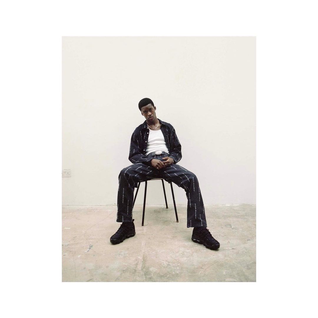 Black man seated on a metal chair against a plain white studio background wearing patterned dark clothing and sneakers