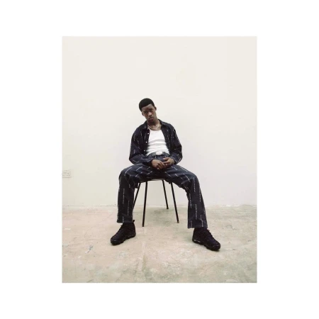 Black man seated on a metal chair against a plain white studio background wearing patterned dark clothing and sneakers