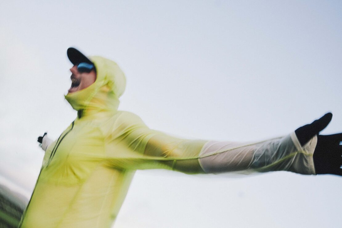 An adult in athletic wear and sunglasses extends their arms in front of a blurred-sky background in a dynamic outdoor shot.