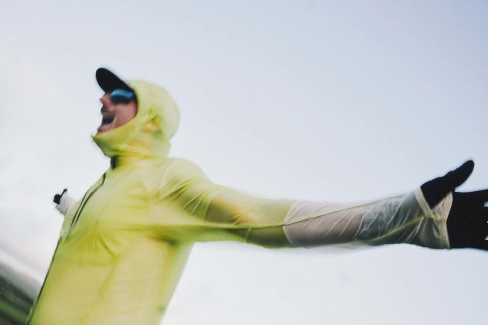An adult in athletic wear and sunglasses extends their arms in front of a blurred-sky background in a dynamic outdoor shot.