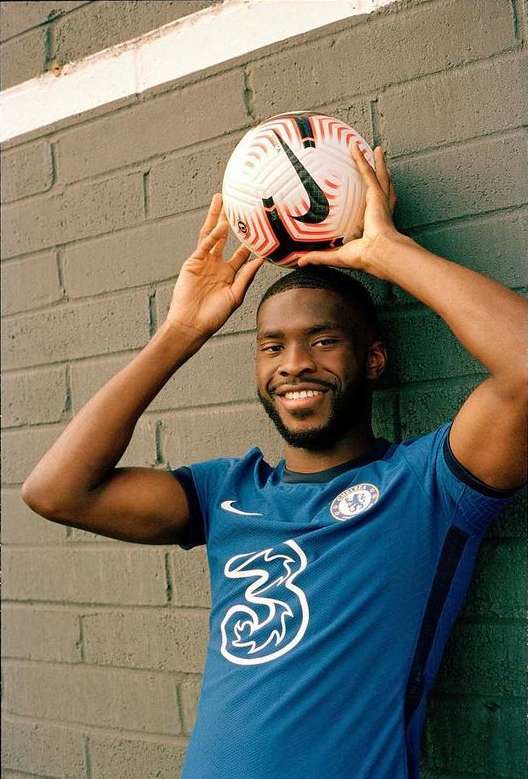 Smiling soccer player poses with a ball on his head wearing a blue Chelsea jersey with the team logo in front of a grey wall.