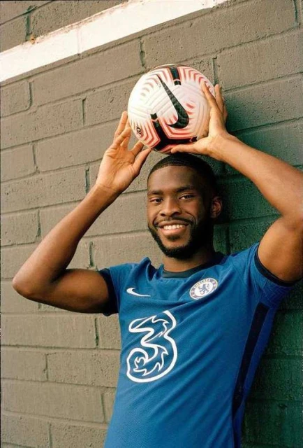 Smiling soccer player poses with a ball on his head wearing a blue Chelsea jersey with the team logo in front of a grey wall.