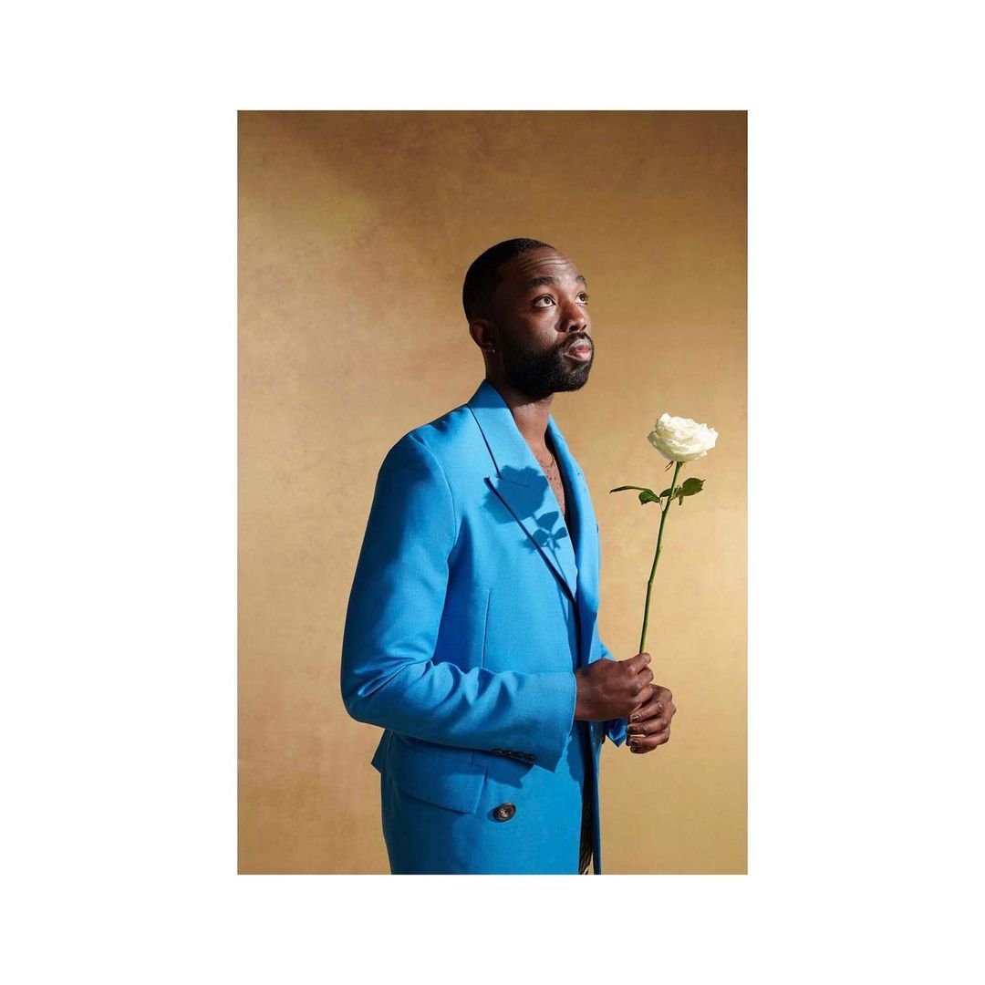Adult man in a bright blue suit and holding a white rose against a textured tan background, looking up, in a studio shot.