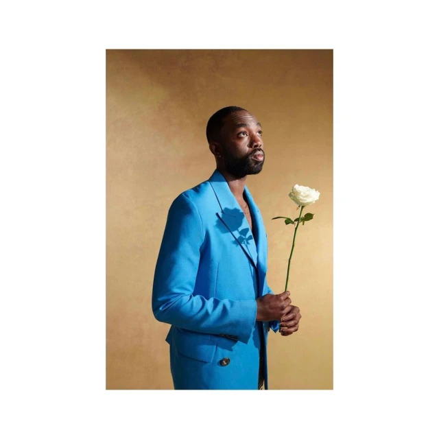 Adult man in a bright blue suit and holding a white rose against a textured tan background, looking up, in a studio shot.