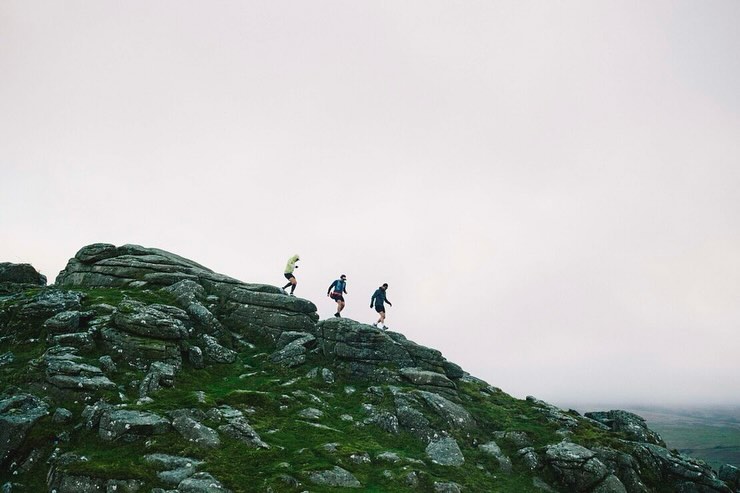 Three people trail running on a rocky, grassy hilltop beneath a cloudy sky, captured in a wide shot emphasizing the natural environment.