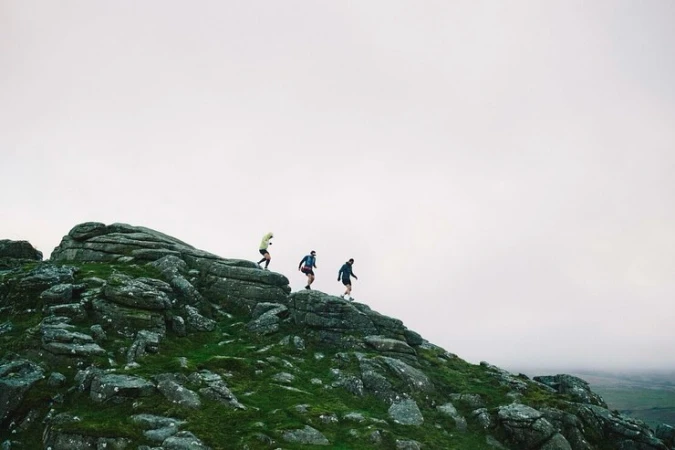 Three people trail running on a rocky, grassy hilltop beneath a cloudy sky, captured in a wide shot emphasizing the natural environment.
