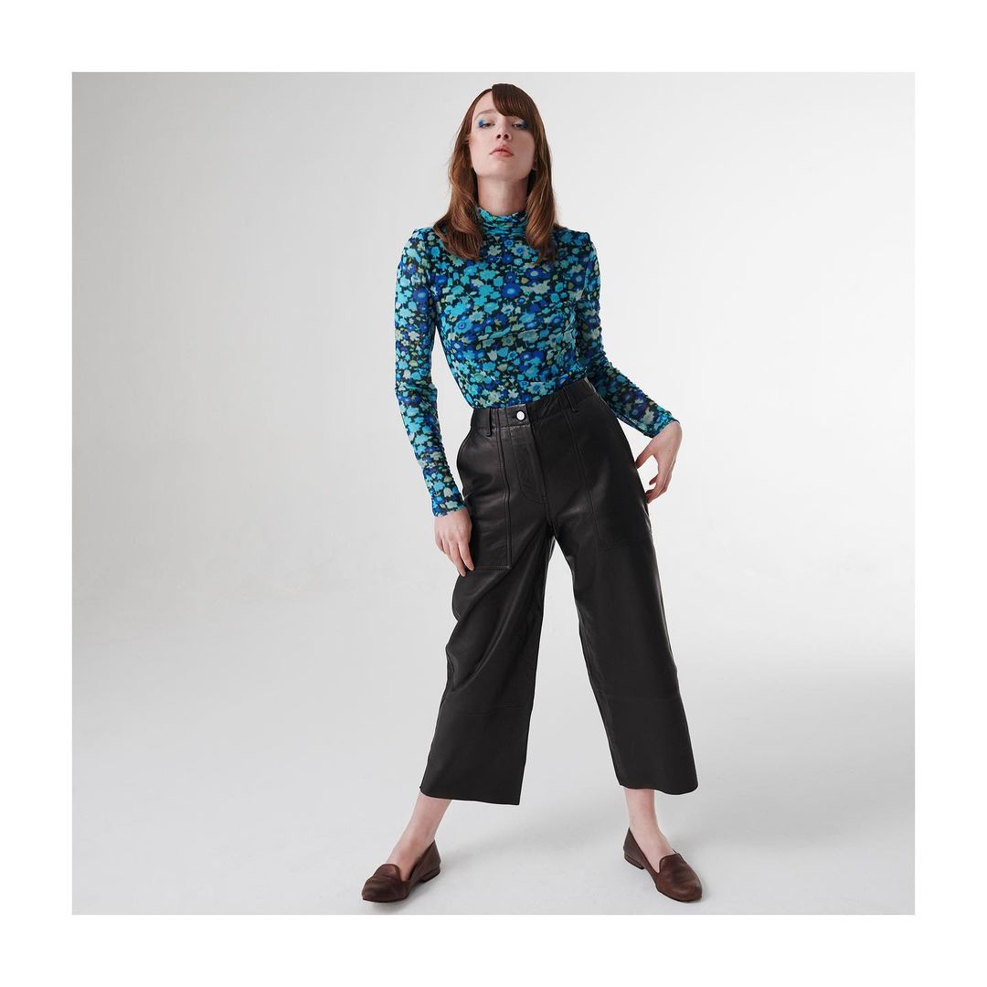 Model wearing a blue floral turtleneck and black leather cropped trousers against a white seamless studio backdrop.