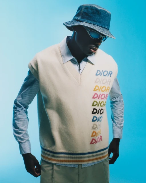 Man in denim bucket hat, striped shirt and sweater vest with colorful DIOR logos, posed in a medium shot against a cyan backdrop.