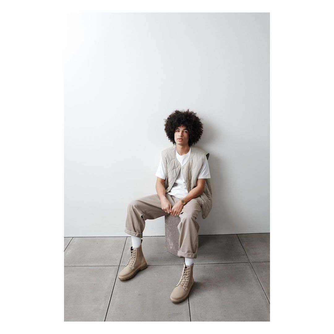Man in vest and boots sitting in a studio. He has dark curly hair and is wearing neutral tones against a light background.