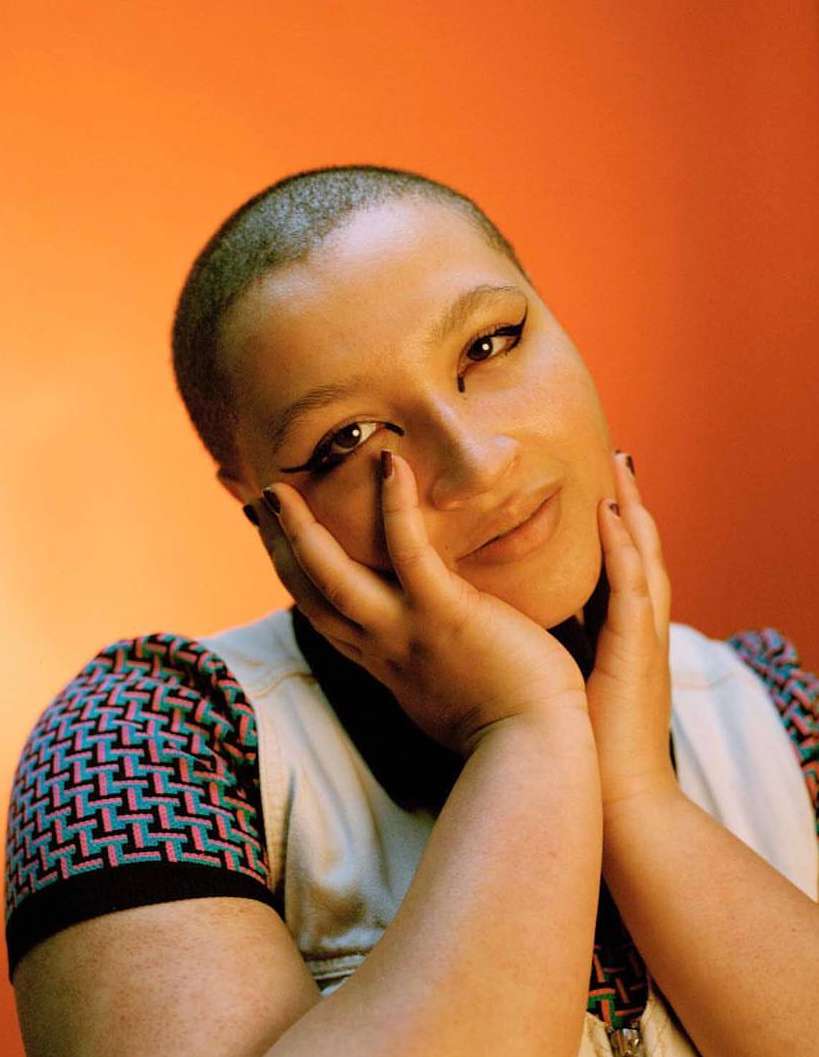 Close-up portrait of a woman with a shaved head resting her face in her hands, against an orange background with soft, diffused lighting.