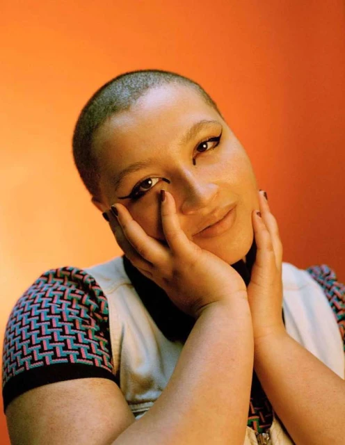 Close-up portrait of a woman with a shaved head resting her face in her hands, against an orange background with soft, diffused lighting.