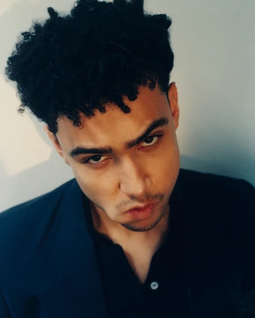Close up portrait of a young man with dark curly hair and a dark jacket looking directly down at the camera