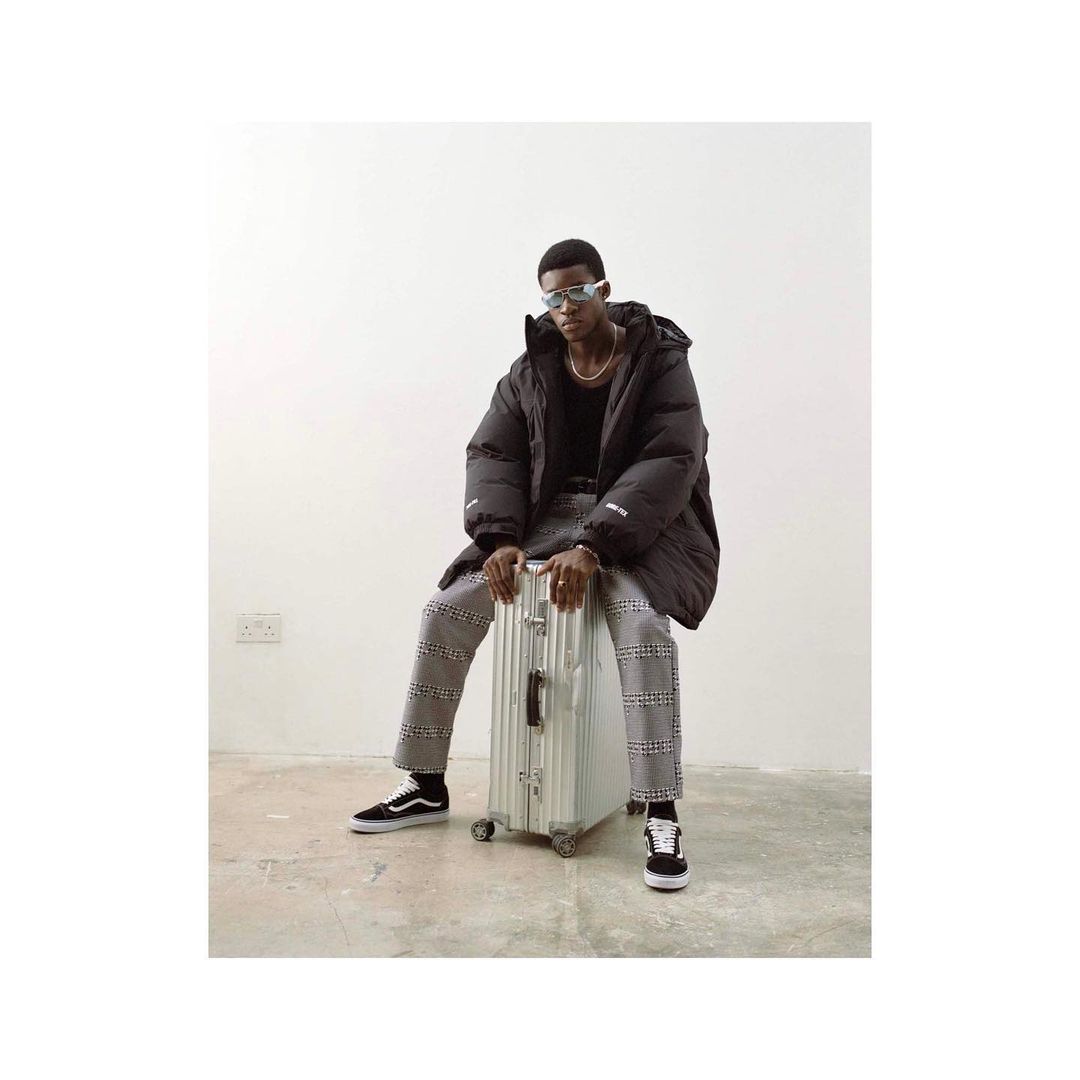 Young Black man wearing a jacket and sunglasses sitting on a silver suitcase indoors against a white wall, wearing checkered pants and sneakers.